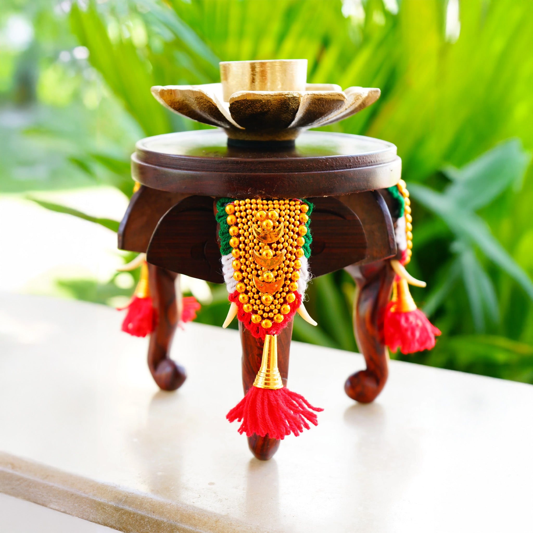 A three-headed wooden elephant stool with carvings and decorative elements, featuring a central golden seat, red hanging embellishments, and intricate woodwork, set against a blurred green background.