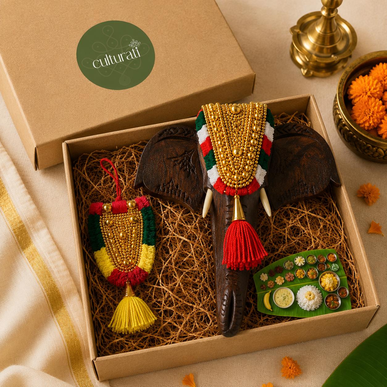 A curated gift box set from Culturati featuring a carved dark brown elephant ornament decorated with a red, green, white, and gold beaded crown, a matching beaded hanging ornament, and a small plate of traditional sweets, all nestled in brown paper straw inside a kraft box.